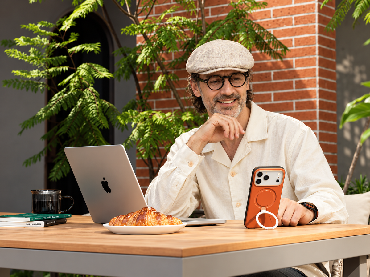 Mann mit Kappe arbeitet im Café am Notebook, orangefarbenes iPhone liegt daneben