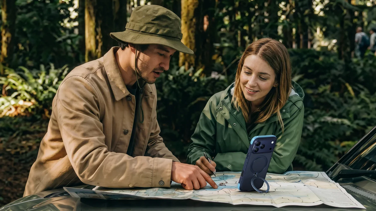 Zwei Personen planen mit einer Landkarte, das Smartphone steht im Ständer auf dem Tisch
