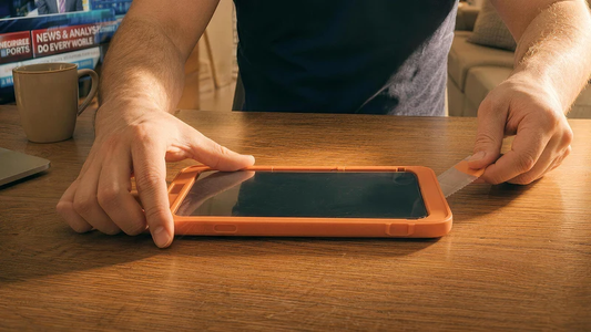 Hände ziehen eine Panzerglasfolie mit einem Rakel auf ein iPhone im orangen Case