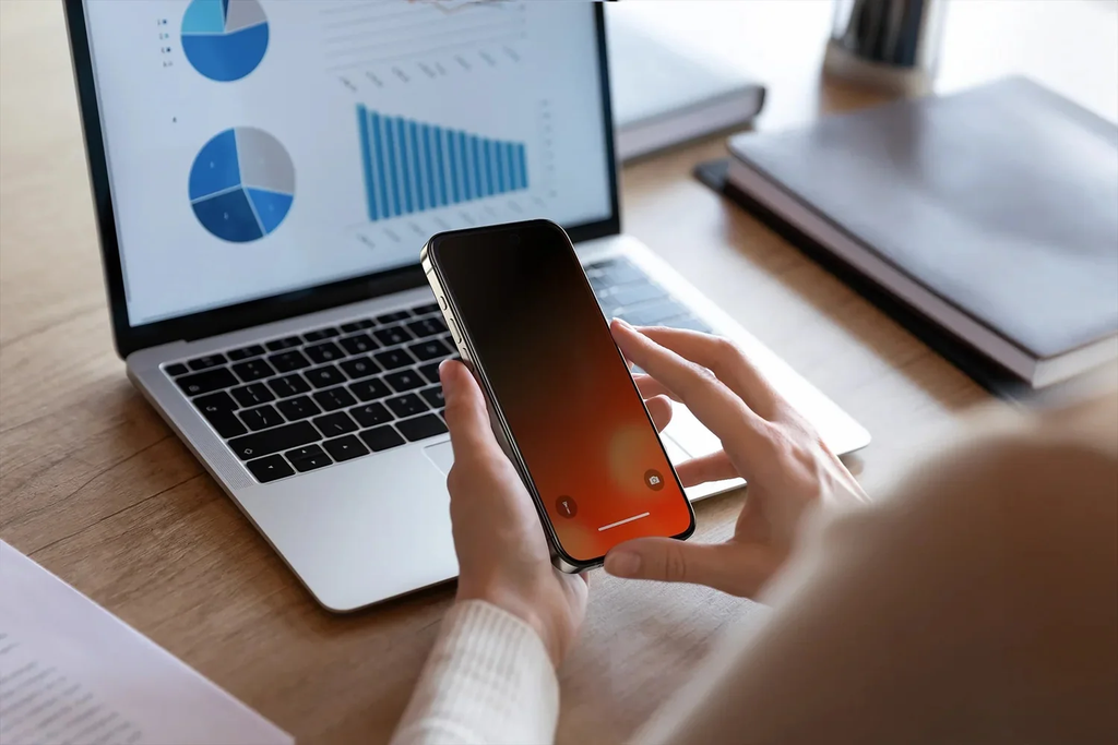 Hände halten ein iPhone mit orangefarbenem Display neben einem Laptop mit Diagrammen