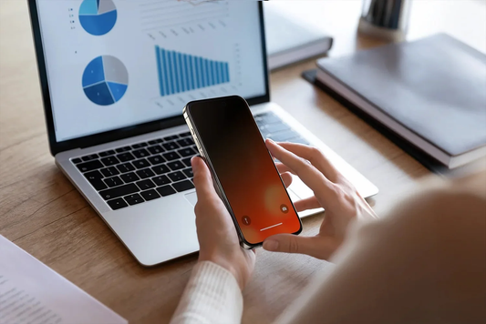 Hände halten ein iPhone mit orangefarbenem Display neben einem Laptop mit Diagrammen