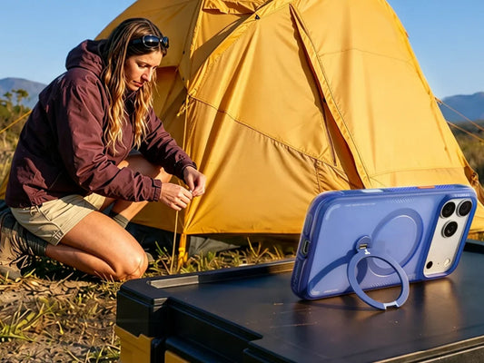 Frau nutzt iPhone mit blauer Hülle am Campingtisch vor Zelt und Bergen